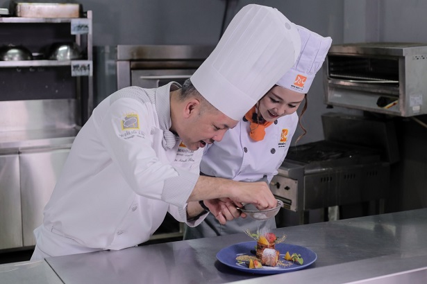Ribi Sachi with a professional chef both wearing a chef uniform while garnishing a food on a blue plate